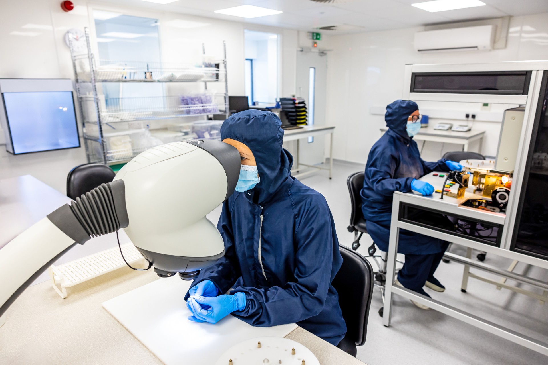 Meridian Medical Cleanroom technician sits at a desk in blue PPE while using machine, another technician sits behind
